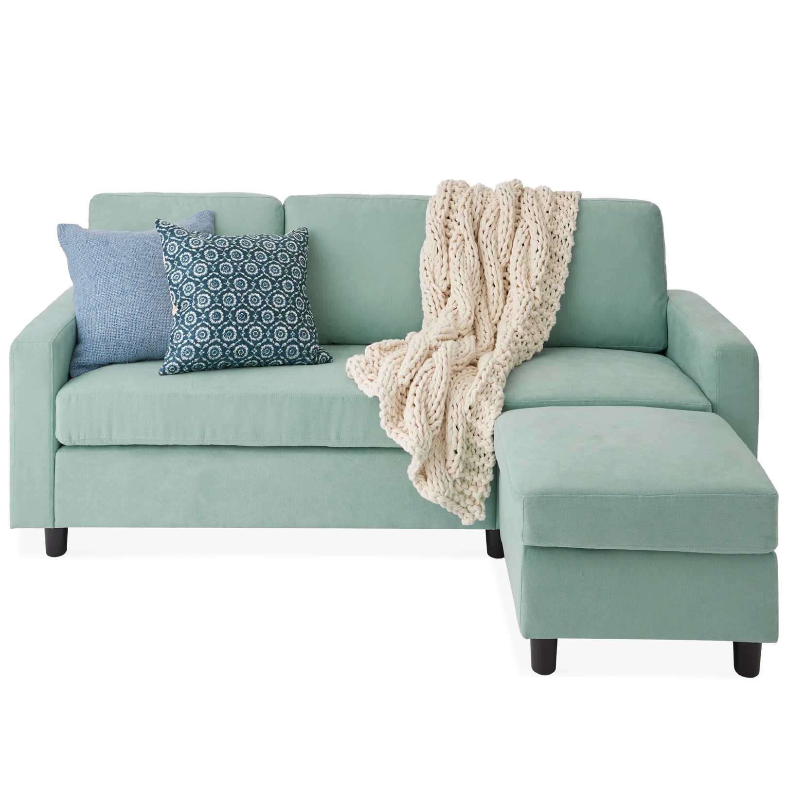 An L-shaped mint green sofa with a chaise, adorned with two blue decorative pillows and a cream chunky knit throw blanket.
