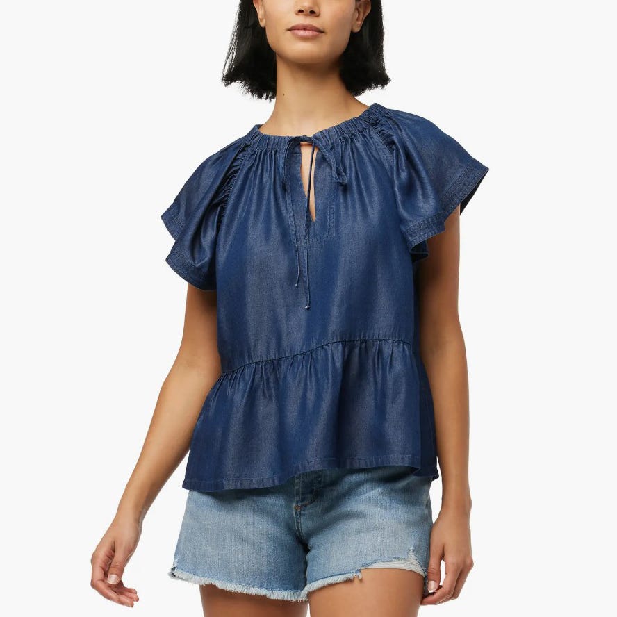 A woman wearing a blue peplum top with gathered sleeves and a keyhole neckline, paired with denim shorts.