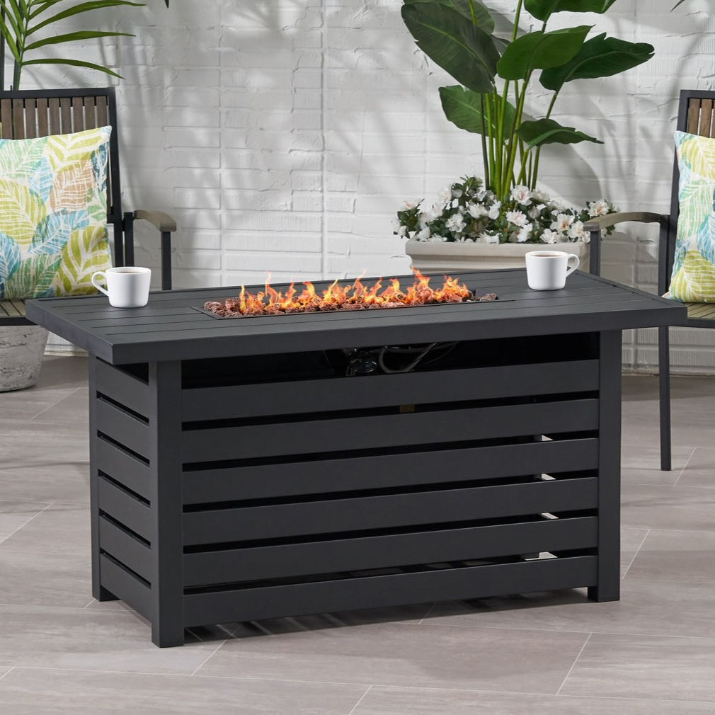 Outdoor patio scene featuring a rectangular black fire pit table with open flames and two chairs with colorful leaf-patterned cushions, surrounded by green and white plants.