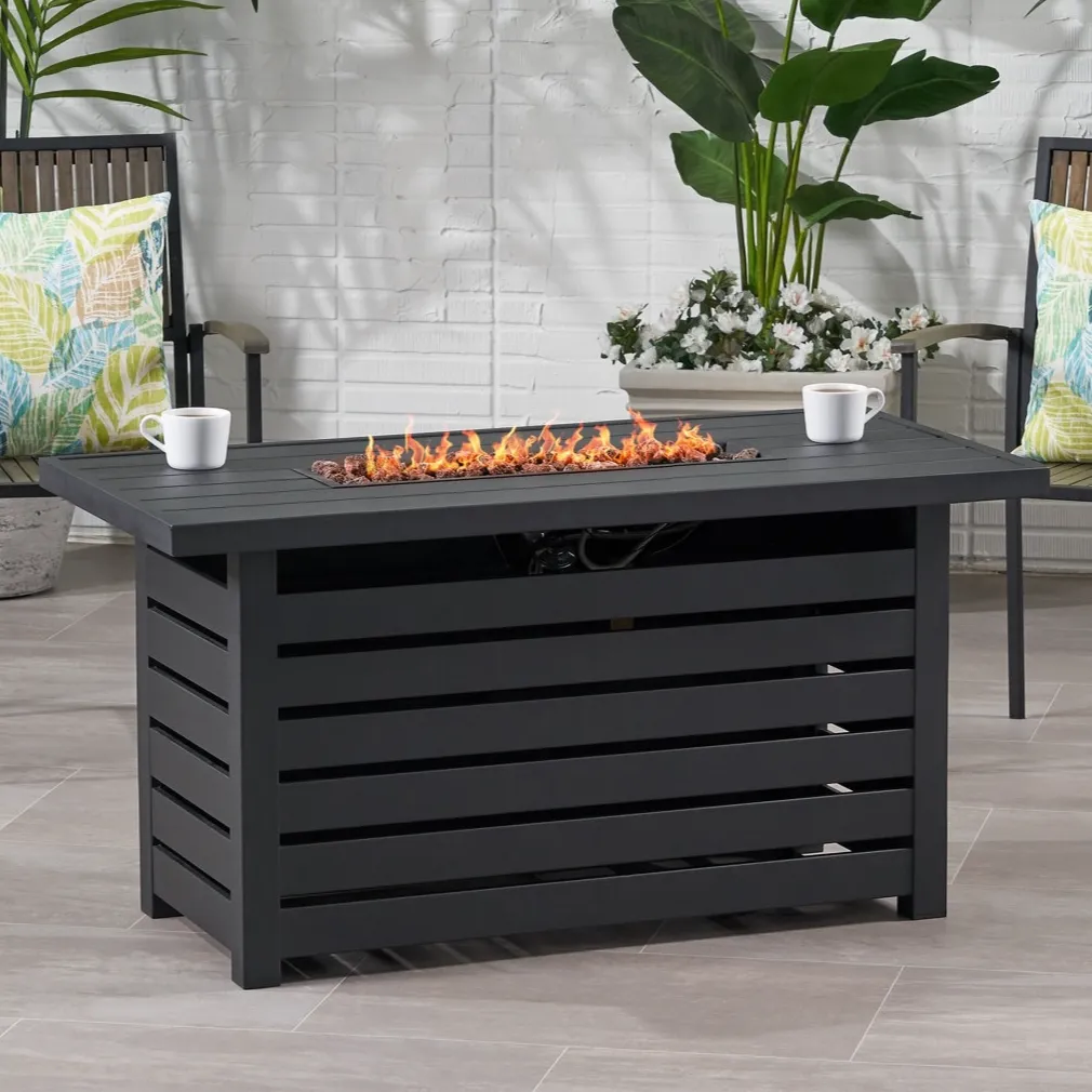 Outdoor patio scene featuring a rectangular black fire pit table with open flames and two chairs with colorful leaf-patterned cushions, surrounded by green and white plants.