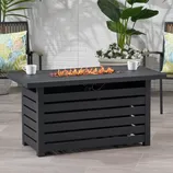 Outdoor patio scene featuring a rectangular black fire pit table with open flames and two chairs with colorful leaf-patterned cushions, surrounded by green and white plants.