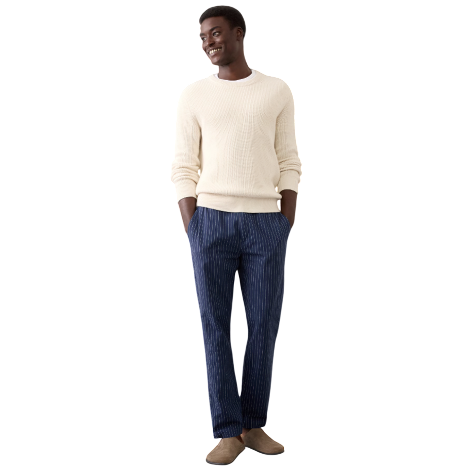 A man is wearing a cream sweater, blue pinstripe pants, and beige slip-on shoes, standing against a light-colored background.