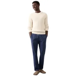 A man is wearing a cream sweater, blue pinstripe pants, and beige slip-on shoes, standing against a light-colored background.