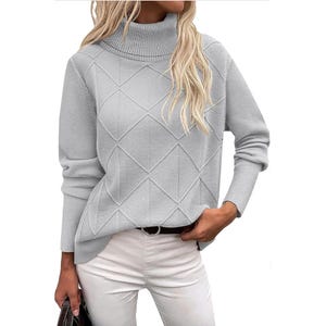 A woman is wearing a light gray knit sweater with a diamond pattern and turtleneck, paired with white pants and holding a handbag.