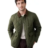 A man wears a quilted green jacket with a brown collar, featuring a zip and snap button closure, along with flap pockets. He pairs it with a light shirt and dark pants.