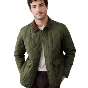 A man wears a quilted green jacket with a brown collar, featuring a zip and snap button closure, along with flap pockets. He pairs it with a light shirt and dark pants.