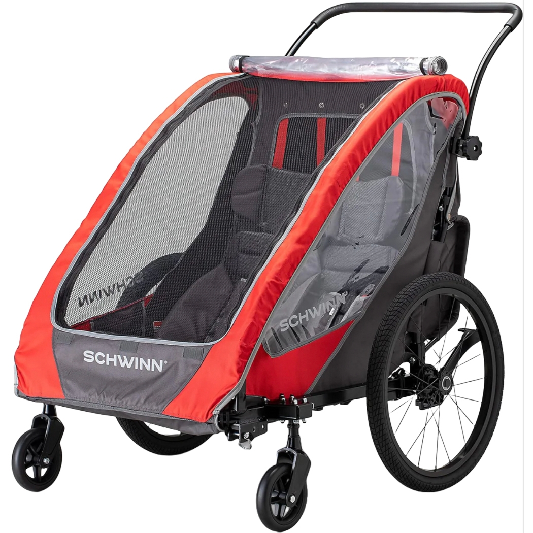 The Schwinn Summit Deluxe Infant Trailer is a red and gray two-seater bike trailer with a mesh front cover and clear side panels. It features a robust frame, large rear wheels, and swiveling front wheels for easy maneuverability.