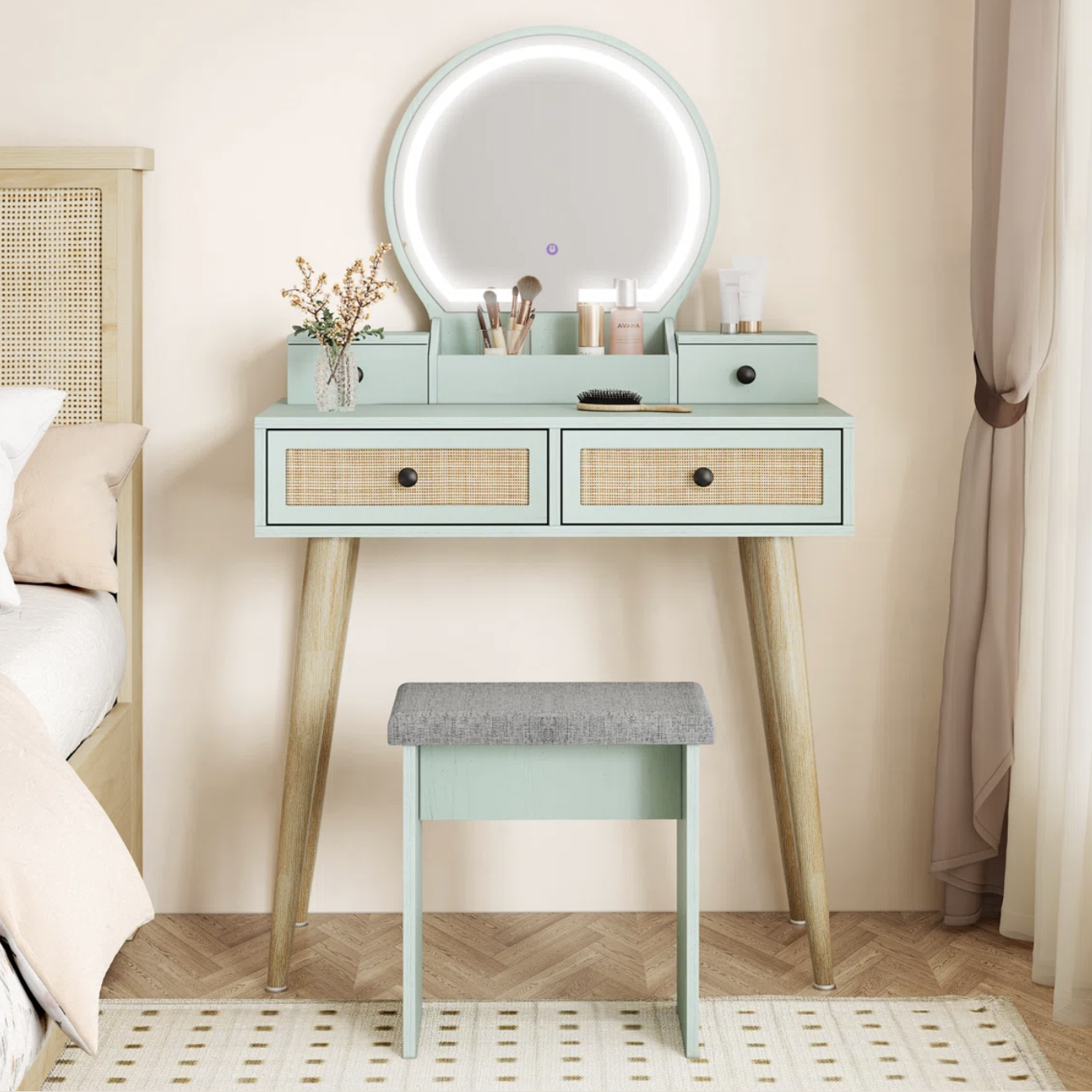 A minimalist vanity set featuring a mint green desk with two rattan-front drawers, a round LED mirror, and a matching stool. The desk surface holds makeup brushes, skincare products, and a small vase with flowers.