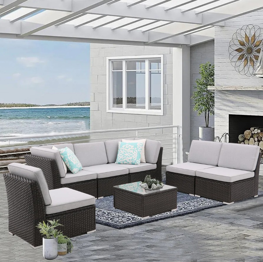 7-piece cushioned patio set includes dark wicker furniture with light gray cushions, featuring a sectional sofa, two single chairs, and a glass-top coffee table, accented with blue patterned throw pillows, set in an outdoor space by the sea.