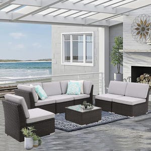 7-piece cushioned patio set includes dark wicker furniture with light gray cushions, featuring a sectional sofa, two single chairs, and a glass-top coffee table, accented with blue patterned throw pillows, set in an outdoor space by the sea.