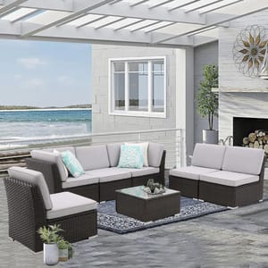7-piece cushioned patio set includes dark wicker furniture with light gray cushions, featuring a sectional sofa, two single chairs, and a glass-top coffee table, accented with blue patterned throw pillows, set in an outdoor space by the sea.