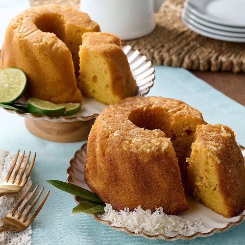 Two Bundt-style rum cakes, each in a 16oz size, are presented on separate plates. One is garnished with lime slices, while the other is topped with shredded coconut.