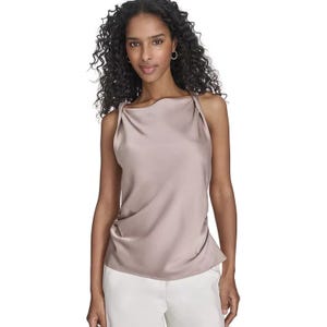 A woman wears a sleeveless Calvin Klein top featuring twisted straps, a soft beige color, and a relaxed drape. The top is paired with white pants.