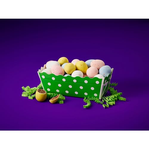 A green dot-patterned paper basket holds pastel-colored, speckled chocolate eggs with cracked open eggs revealing chocolate filling, set on green paper grass against a purple background.