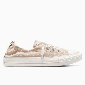A low-top Converse Chuck Taylor sneaker in light beige with white polka dots, featuring a white rubber toe cap, white laces, and a white midsole.