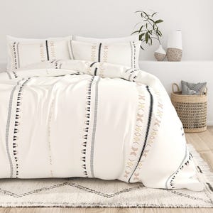 A white bedding set features geometric patterns in black and pale gold, resting on a patterned rug. Nearby, a plant in a vase and a woven basket with pillows complete the décor.