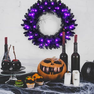 Halloween-themed setup with a carved pumpkin, purple-lit wreath, black and red candles in bottles, black apples, and festive cupcakes on a black cloth with cobwebs.