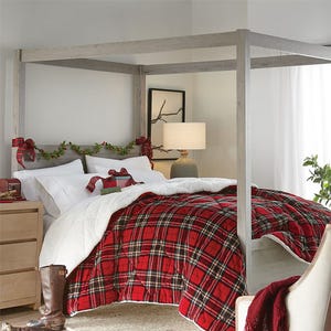 A cozy bedroom features a canopy bed with a red plaid blanket, white bedding, and Christmas decorations. A bedside table holds a lamp and is flanked by brown boots on the floor.
