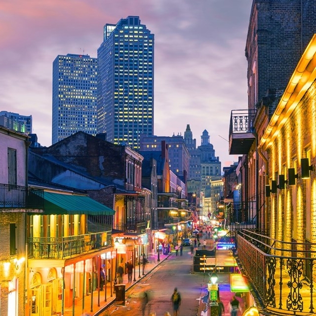Bustling evening street scene with colorful lights and historic buildings, likely in a vibrant urban area with skyscrapers in the background.