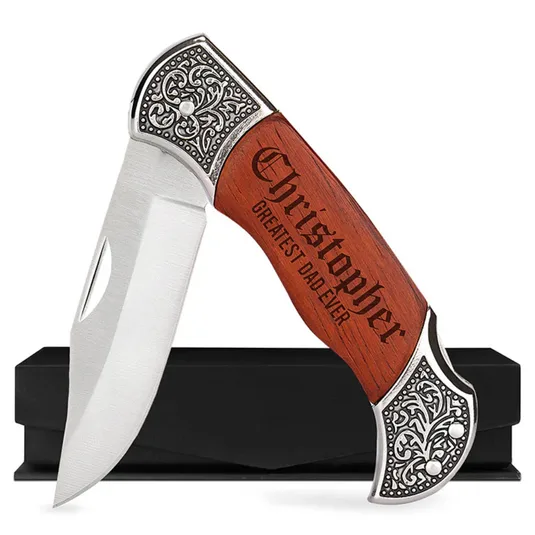 A folding knife features a wooden handle engraved with \“Christopher Greatest Dad Ever\“ and ornate metal detailing, shown against a black box.