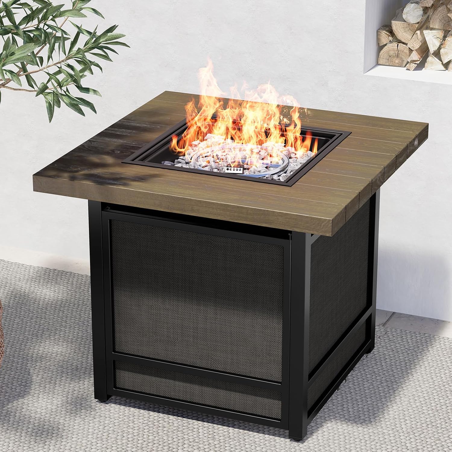 A square fire pit table with a wood finish top and black metal base, displaying a lit fire with glass rocks surrounding the flames.