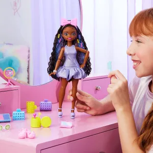 A doll with long, curly hair in a purple dress and pink bow stands on a pink vanity, surrounded by colorful toy beauty accessories, including a mirror, brush, makeup, and shoes. A child interacts with the doll, holding a small comb.