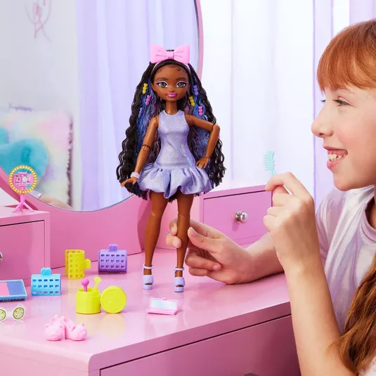 A doll with long, curly hair in a purple dress and pink bow stands on a pink vanity, surrounded by colorful toy beauty accessories, including a mirror, brush, makeup, and shoes. A child interacts with the doll, holding a small comb.