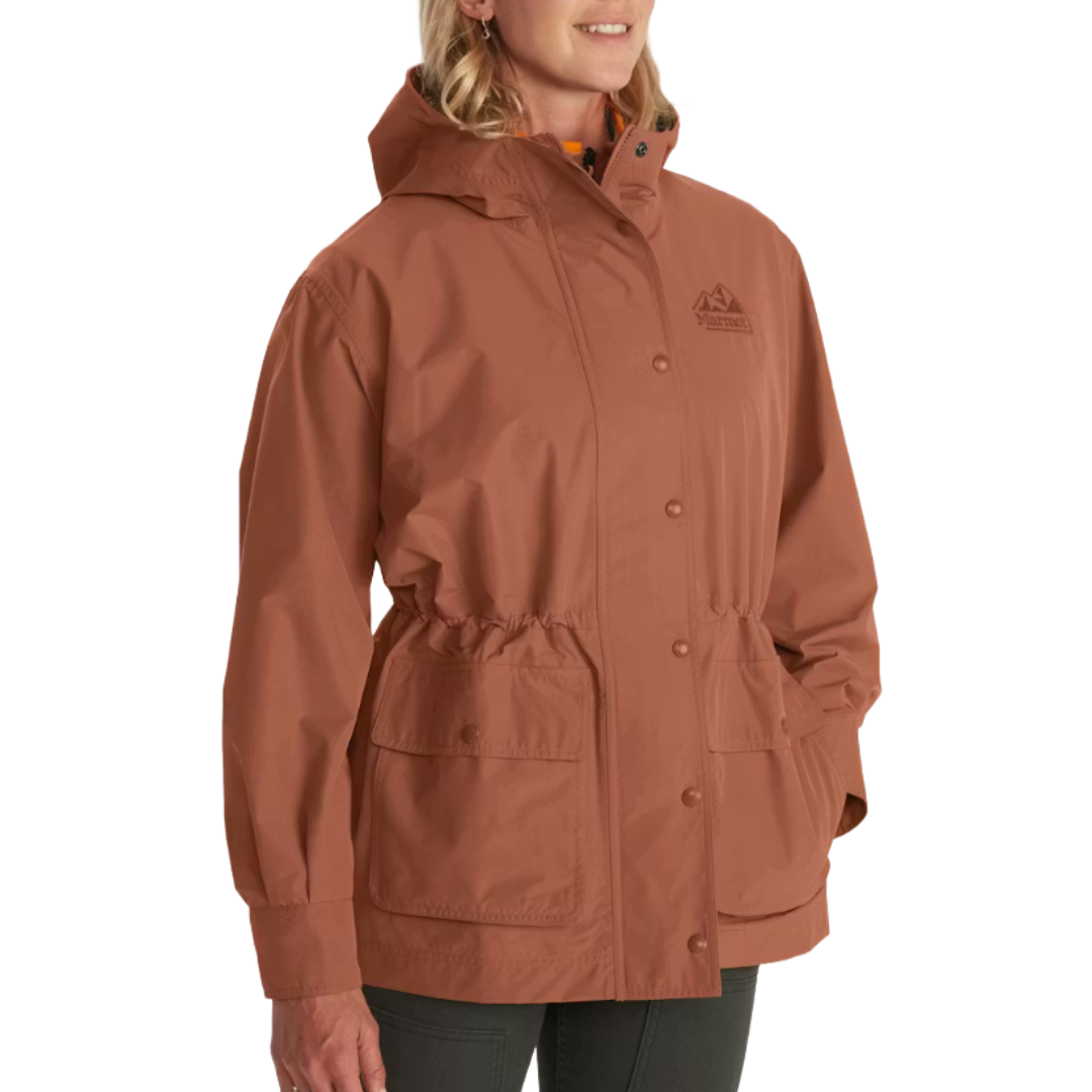 A woman is wearing a rust-colored hooded rain jacket with a drawstring waist and two large front pockets.