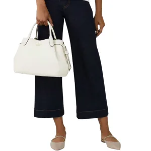 A person is holding a white handbag while wearing dark wide-legged jeans and beige flats with a rhinestone strap.