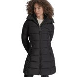 A woman is wearing a long black puffer coat with a front zipper, side pockets, and a high collar, over a light-colored sweater.