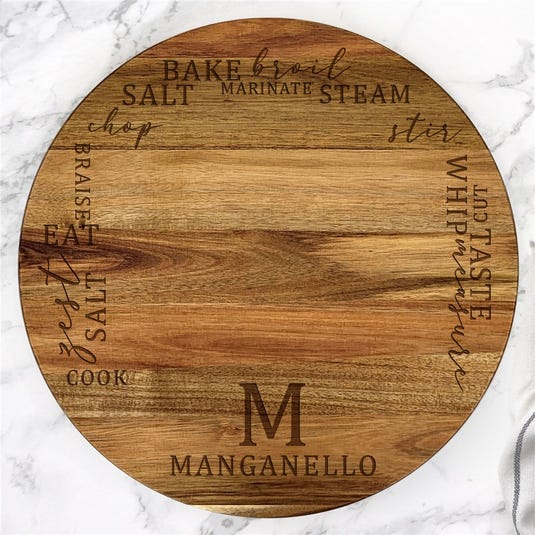 A round wooden Lazy Susan with various cooking-related words like \“bake,\“ \“broil,\“ and \“stir\“ engraved along the edge, featuring a large \“M\“ and the name \“Manganello\“ at the center.