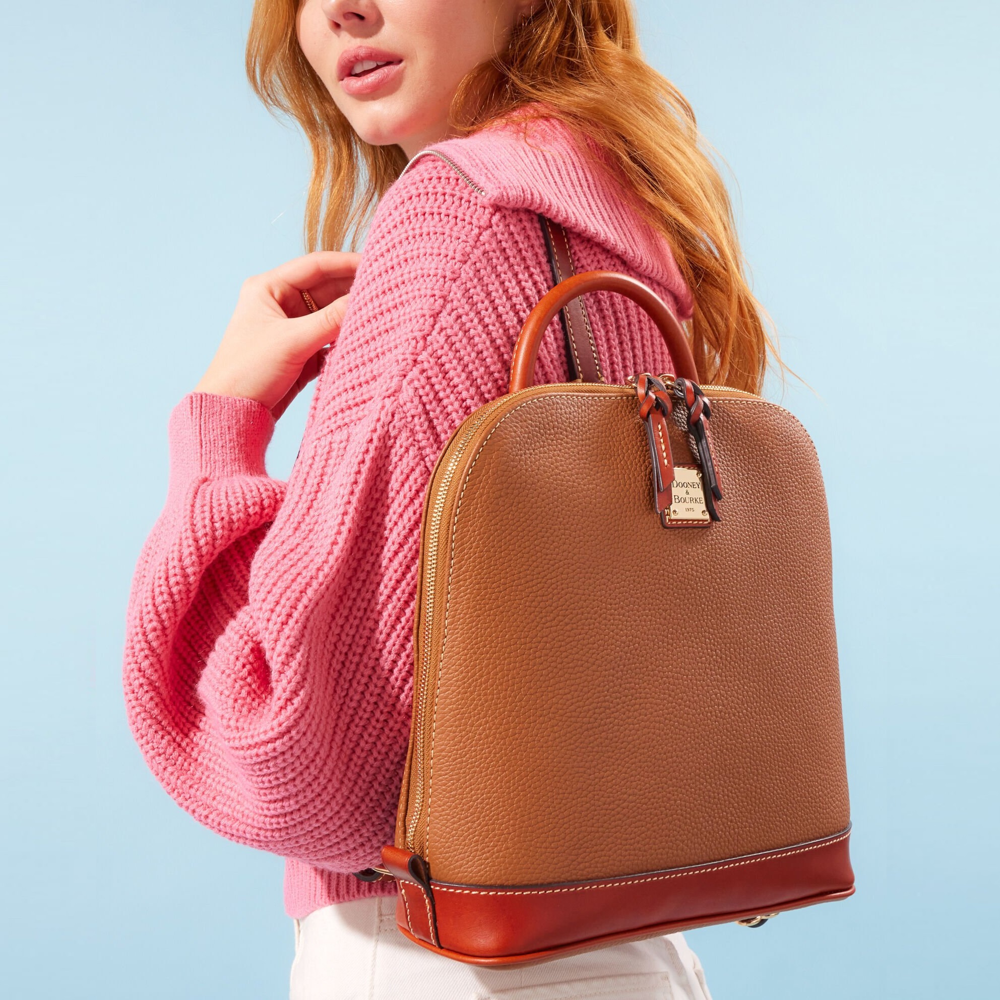 A woman wearing a pink knitted sweater carrying a brown leather backpack with tan accents.