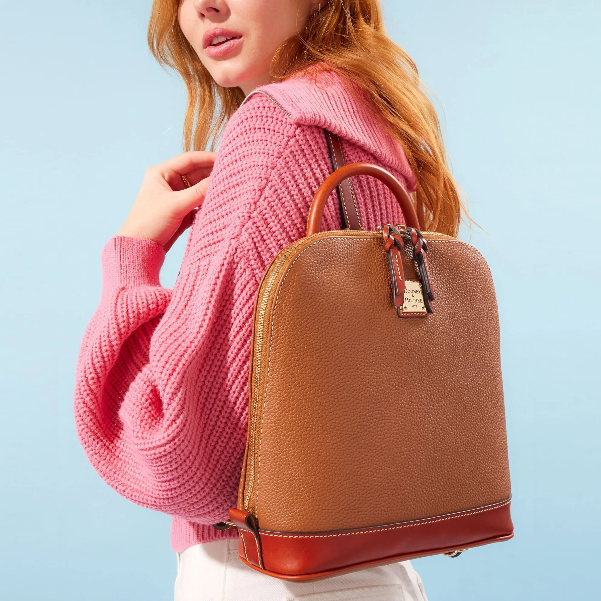 A woman wearing a pink knitted sweater carrying a brown leather backpack with tan accents.