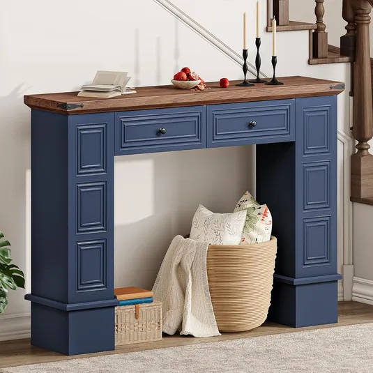 Blue console table with a wooden top featuring two drawers, positioned in a hallway with decorative items on top, including candlesticks and a bowl of fruit. Below, it displays a basket filled with pillows and a blanket, alongside a basket and stacked books.