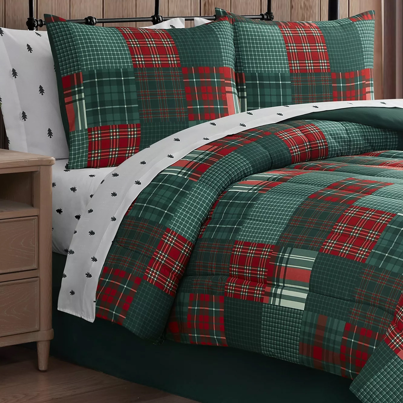 A bedding set features a red and green plaid quilt with matching pillow shams, paired with white sheets adorned with small black tree patterns, set against a wooden background.