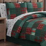 A bedding set features a red and green plaid quilt with matching pillow shams, paired with white sheets adorned with small black tree patterns, set against a wooden background.
