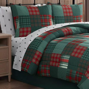 A bedding set features a red and green plaid quilt with matching pillow shams, paired with white sheets adorned with small black tree patterns, set against a wooden background.