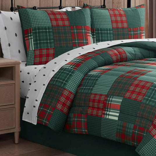 A bedding set features a red and green plaid quilt with matching pillow shams, paired with white sheets adorned with small black tree patterns, set against a wooden background.