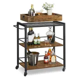 A rolling bar cart with three wooden shelves and a black metal frame holds various bottles, a decanter, ice bucket, and a serving tray with glasses and a potted plant on top.