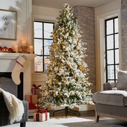 A 7.5-foot flocked Christmas tree with warm white lights stands in a home setting, adorned with a subtle dusting of snow and accompanied by wrapped gifts at its base.