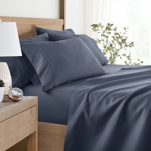 A neatly made bed with navy blue sheets and pillowcases is shown, accompanied by a wooden nightstand with a white lamp, small potted plants, and a clock.