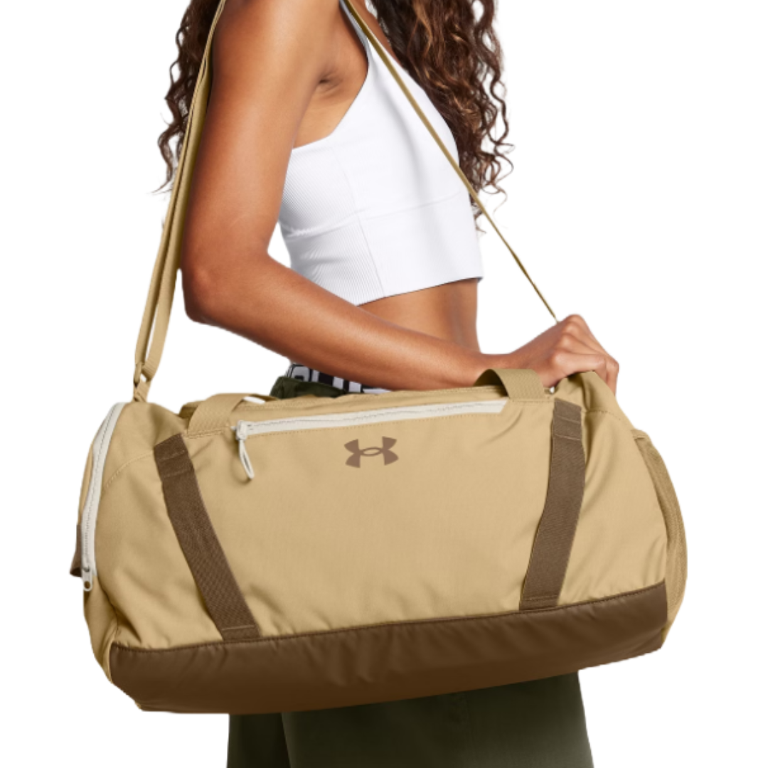 A person carries a tan Under Armour duffel bag with a long shoulder strap and brown accents while wearing a white crop top.