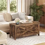 Living room featuring a beige sectional sofa, a rustic wooden chest-style coffee table with an \“X\“ pattern, a white patterned rug, decorative pillows, and a potted plant.