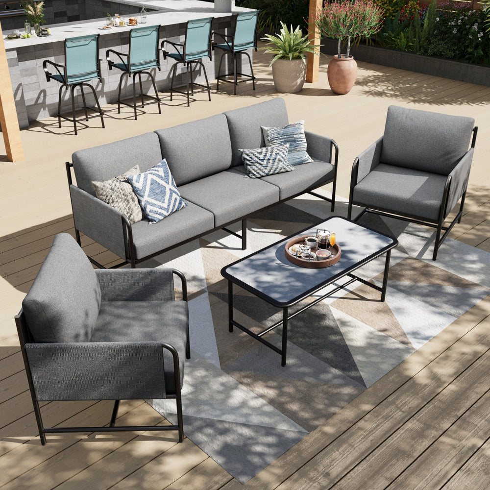 Outdoor patio set with a modern design includes a gray cushioned sofa, two matching armchairs, and a rectangular coffee table on a patterned rug. Four teal bar stools are visible at a nearby outdoor bar area.