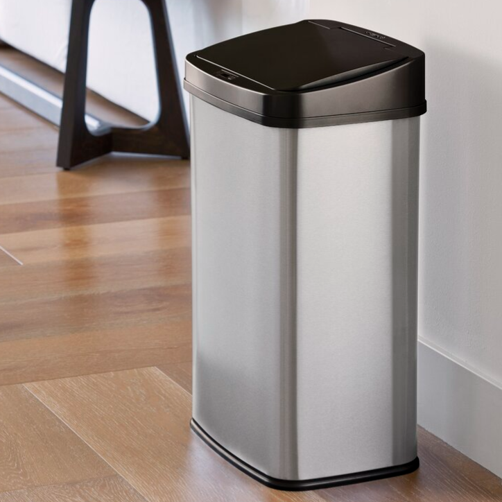 A silver rectangular trash can with a black lid, placed on a wooden floor near a white wall.