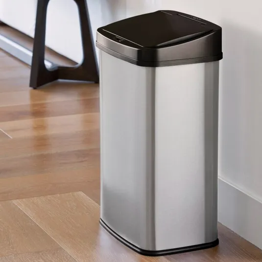 A silver rectangular trash can with a black lid, placed on a wooden floor near a white wall.