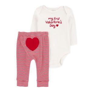 The outfit includes a long-sleeve white onesie with \“my first Valentine's Day\“ written in red, paired with red-and-white striped pants featuring a red heart on the back.