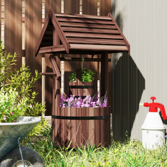 A decorative wooden wishing well with a roof, filled with purple flowers, is situated beside a metal wheelbarrow and near a white garden sprayer with a red handle.