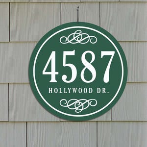 A round green house address plaque with the number 4587 and \“HOLLYWOOD DR.\“ in white letters, featuring decorative scroll designs above and below the text.