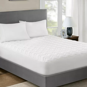 A bed with white fitted sheet, two pillows, and a quilted mattress pad, beside a nightstand with a lamp.